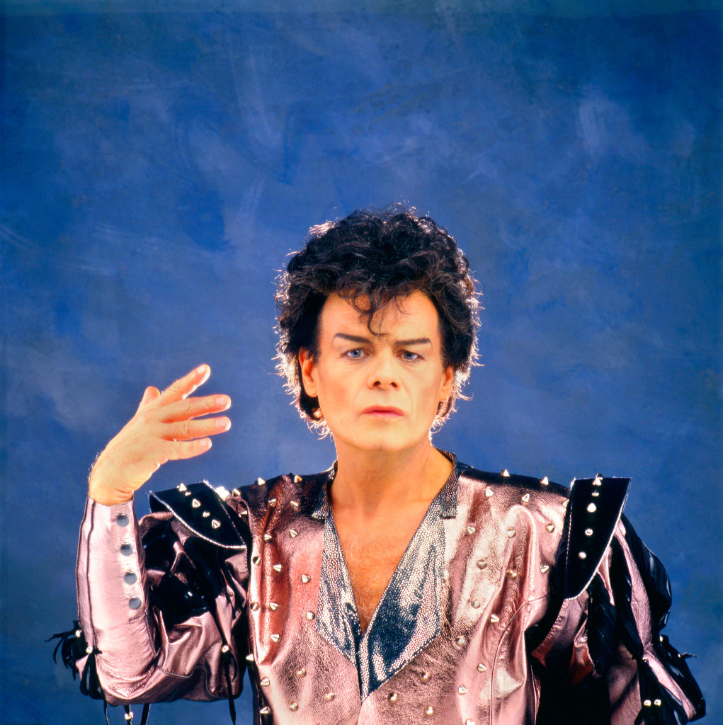 Person with styled hair wears a shiny, studded costume with raised hand, striking a pose against a plain background