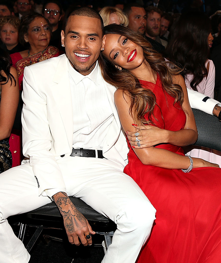 Chris Brown and Rihanna smiling while seated, Chris in a white suit and Rihanna in a red sleeveless dress, leaning her head on his shoulder