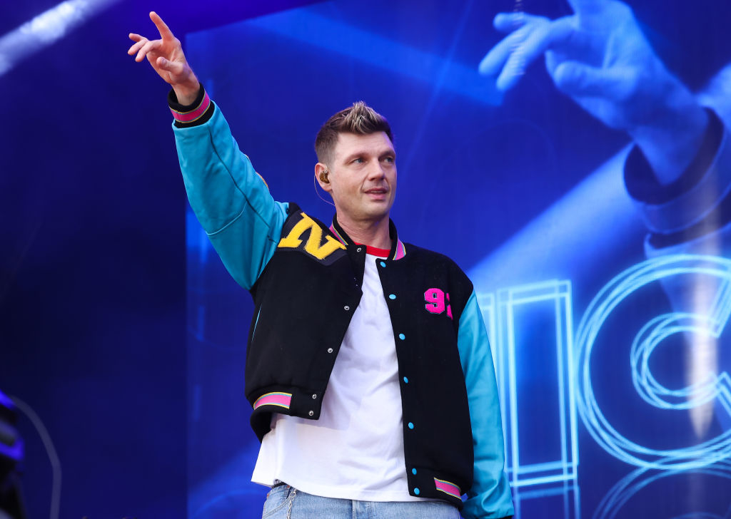 Nick Carter is performing on stage, wearing a varsity jacket and jeans, raising one arm with fingers pointed towards the sky