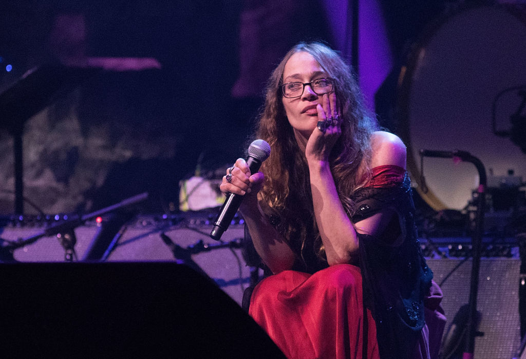 Fiona Apple sits on stage during a performance, holding a microphone and looking thoughtful. She is wearing a flowing, off-shoulder dress. Musical equipment is in the background