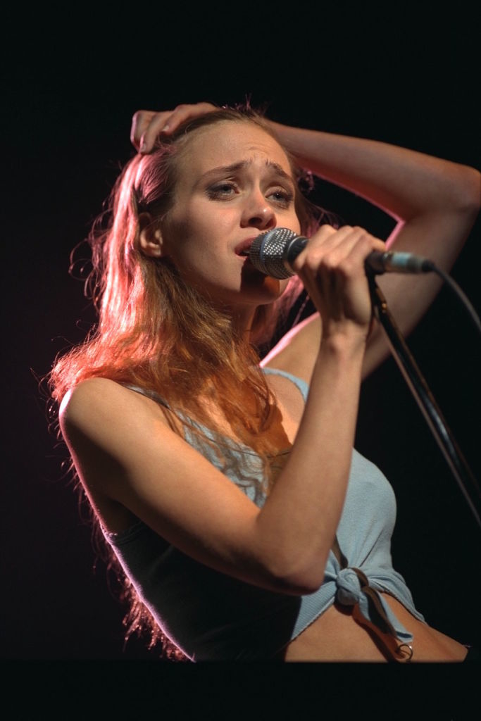 Fiona Apple is on stage, holding a microphone and performing. She has one hand on her head, wearing a sleeveless top