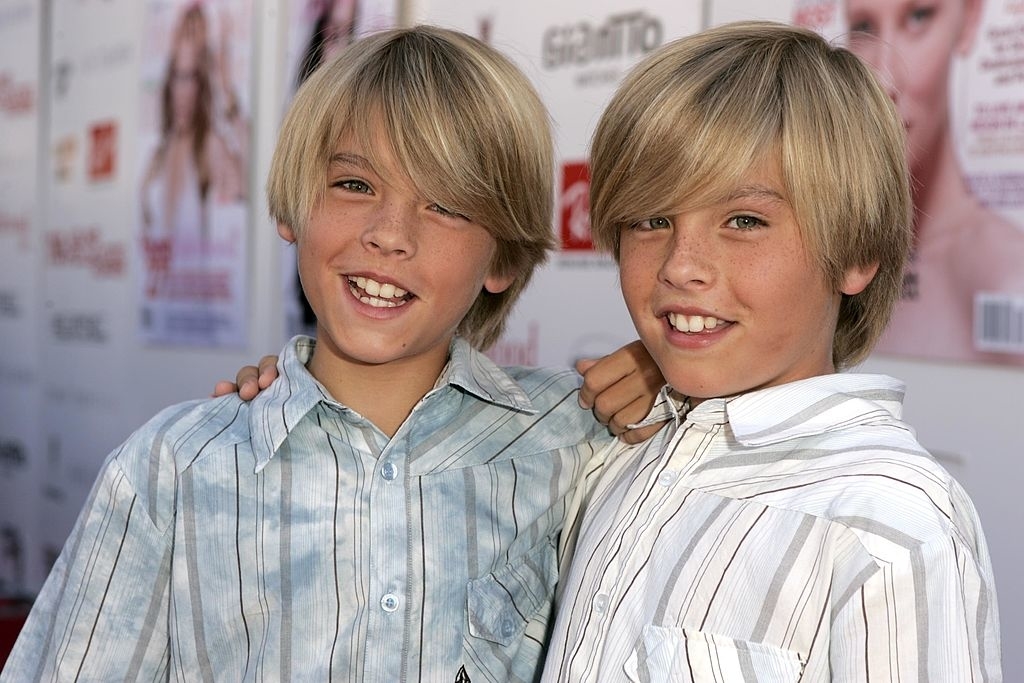 Dylan and Cole Sprouse on the red carpet, smiling with their arms around each other, wearing casual button-up shirts
