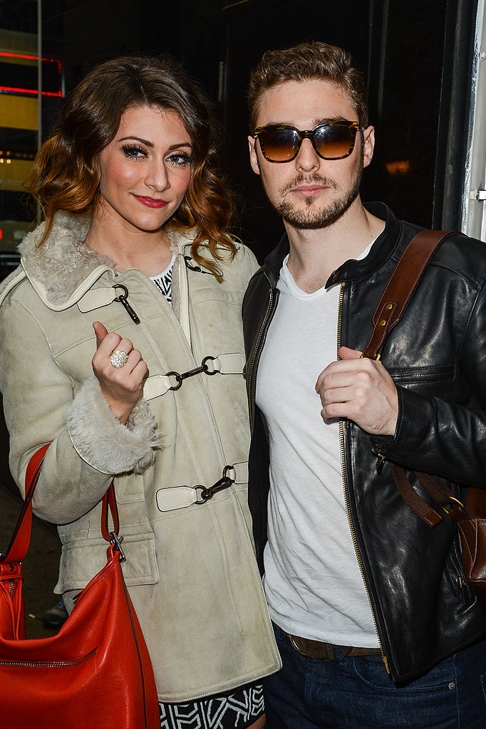 I don't know who these people are. A man and woman are standing, both wearing jackets. The man has sunglasses and a backpack, the woman holds a red handbag