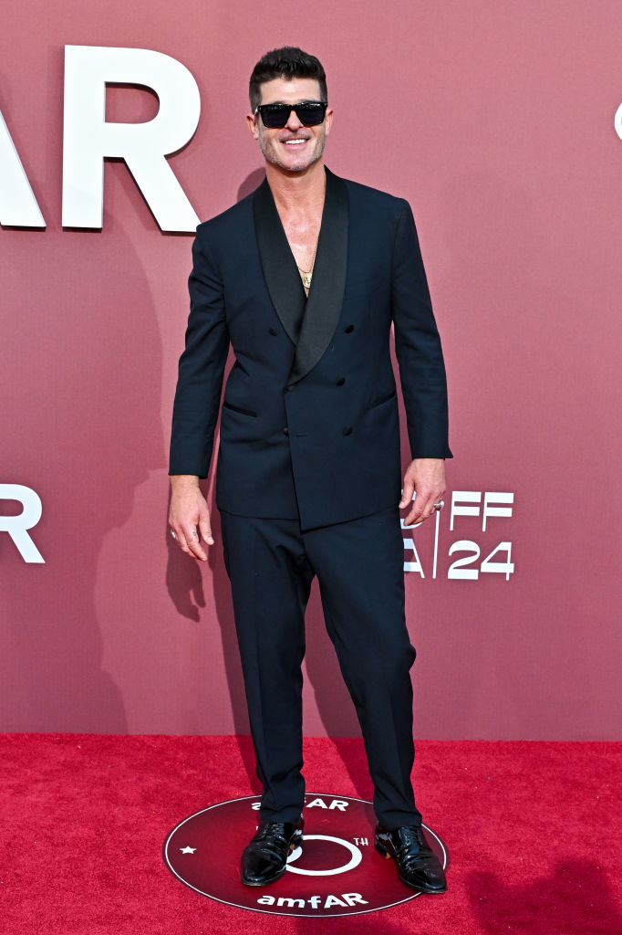 Robin Thicke at an amfAR event, wearing a black tuxedo with a shawl collar and black shoes, smiling and standing on a red carpet