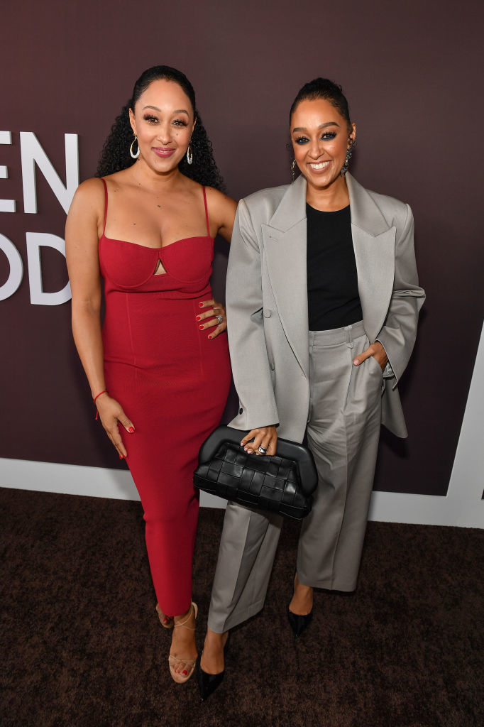 Tamera Mowry in a red dress and Tia Mowry in a grey suit with a black top smile at a red carpet event
