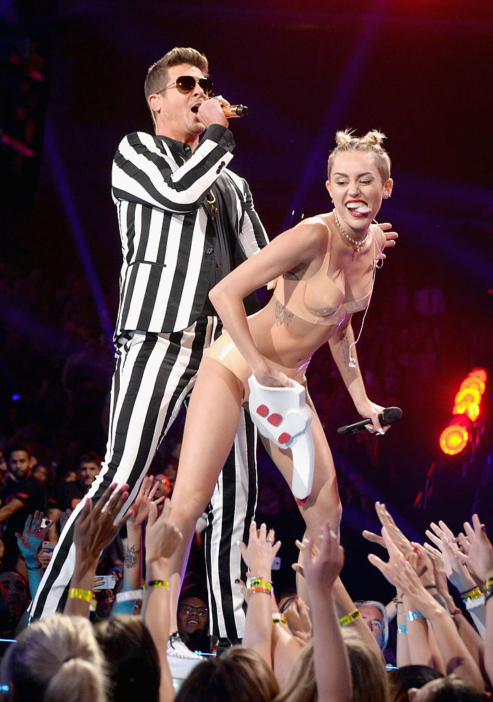 Robin Thicke performs wearing a striped suit while Miley Cyrus dances next to him in a nude outfit holding an oversized foam finger