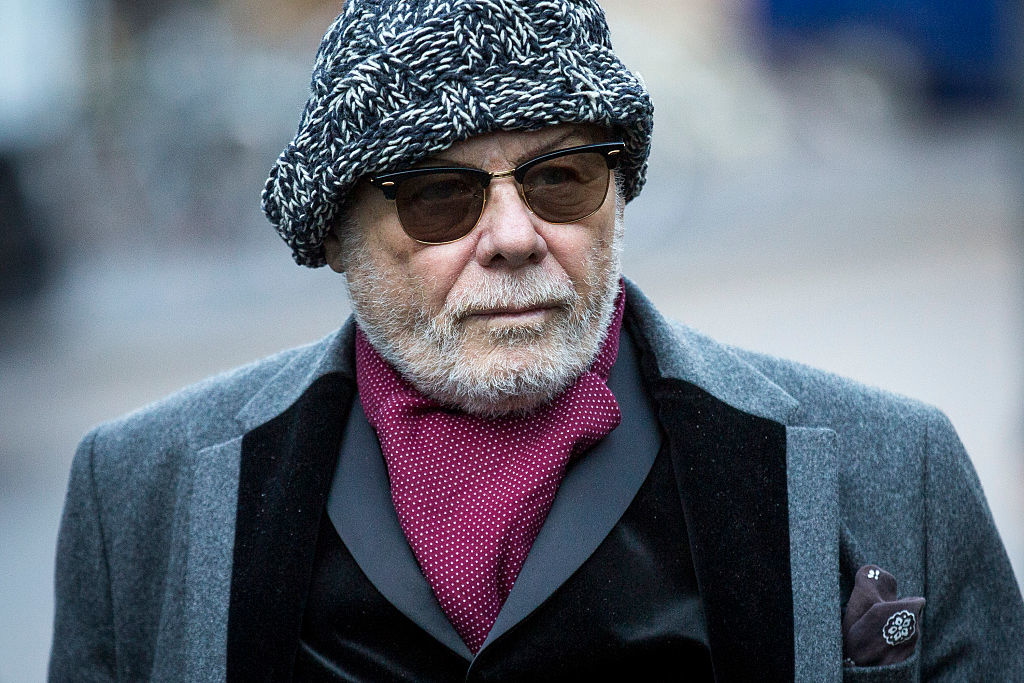An older man with a beard and sunglasses wearing a patterned hat, gray overcoat, black velvet blazer, and a red patterned scarf