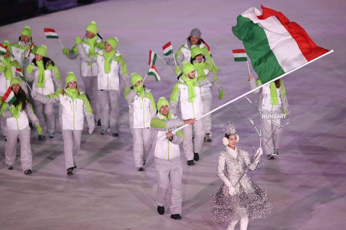 Hungarian athletes march at a sports event, led by a flag bearer holding Hungary’s flag. Participants wear winter sportswear with green hats and wave small flags
