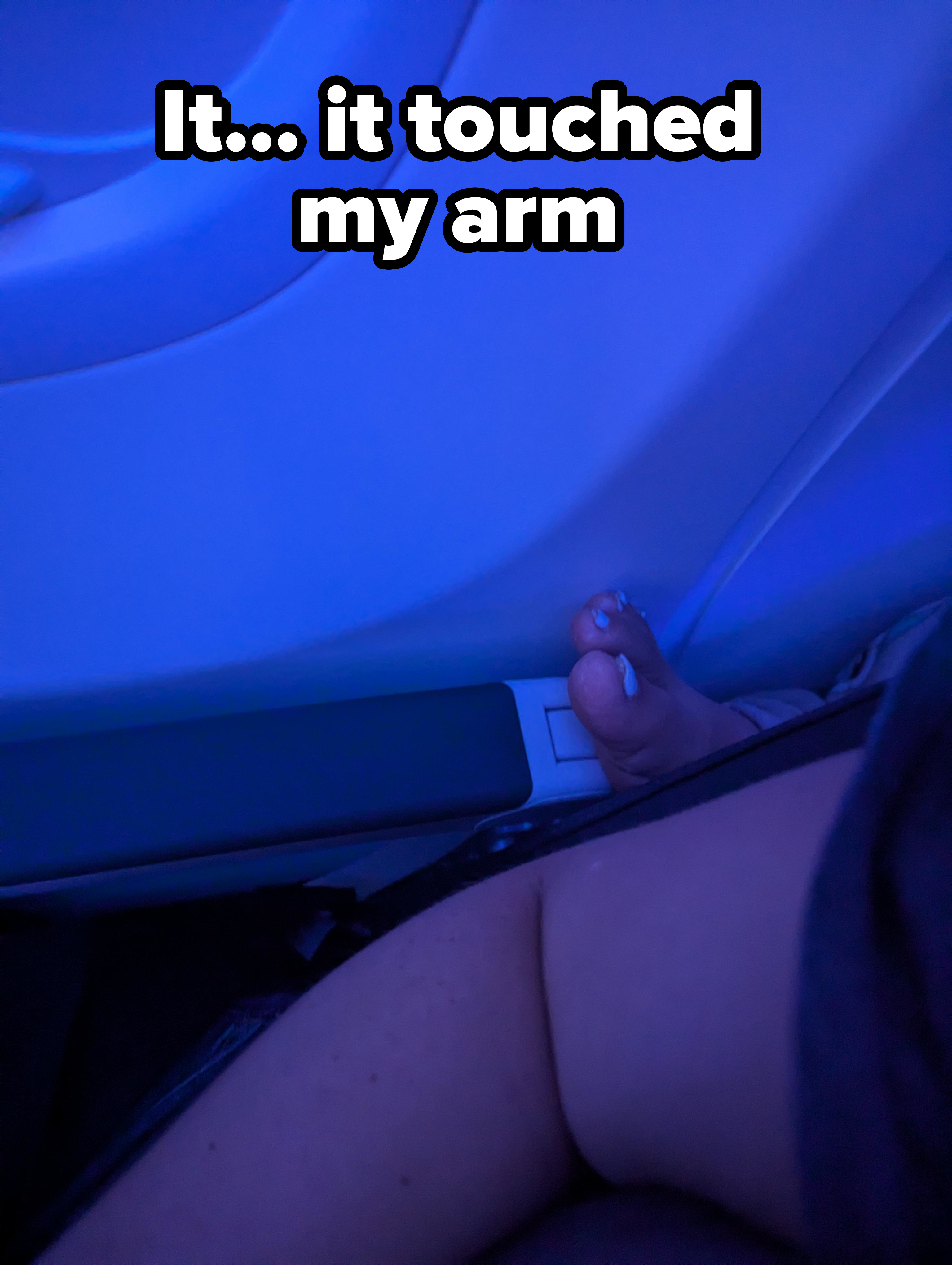 A person's bare foot with blue nail polish is resting against the interior wall of an airplane under the dim cabin light
