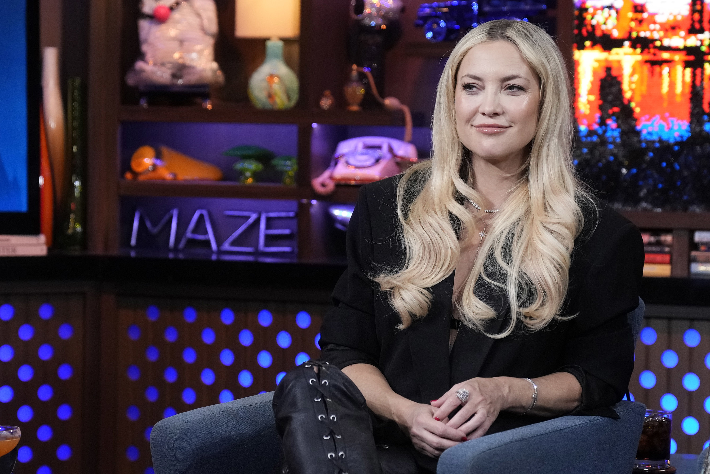 Kate Hudson sits on a talk show set, wearing a stylish black outfit