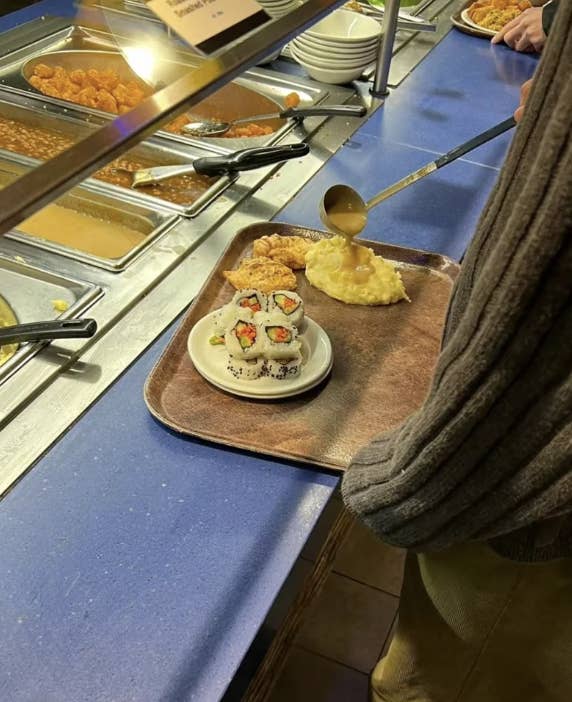 A person is serving food at a buffet, with mashed potatoes, sushi rolls, and fried chicken on their tray