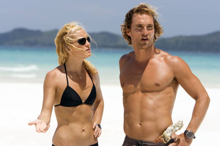 A man and woman on a beach; the woman (in a bikini, wearing sunglasses) gestures, while the man (shirtless, holding a seashell) looks into the distance