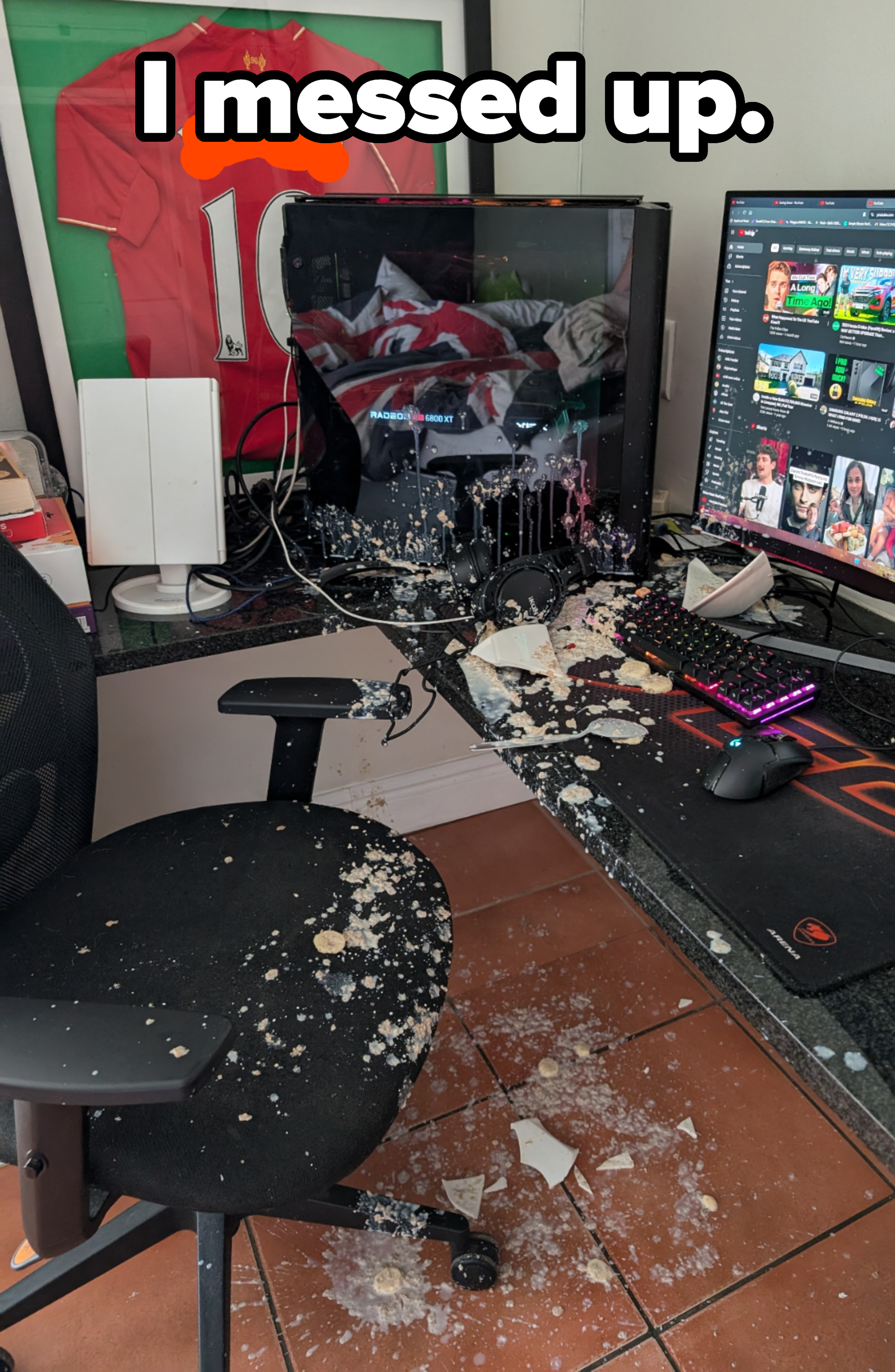 A cluttered desk with a broken monitor, spilled food, and debris. A sports jersey is framed above the desk, and a YouTube page is open on another monitor