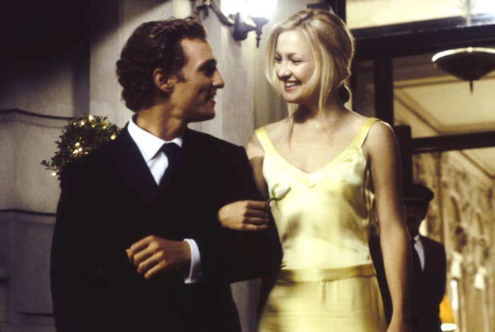 Matthew McConaughey in a suit and Kate Hudson in an elegant dress smiling and walking arm in arm outside a building, accompanied by people in the background