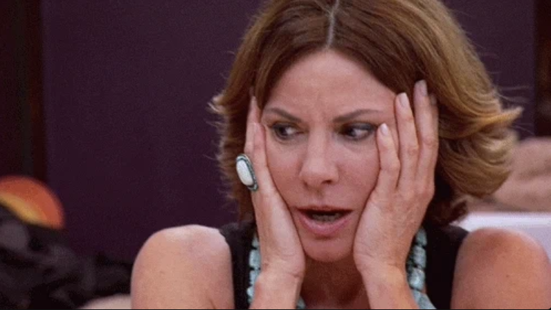 A woman with short brown hair, wearing a black top and turquoise jewelry, holds her face with both hands, looking surprised