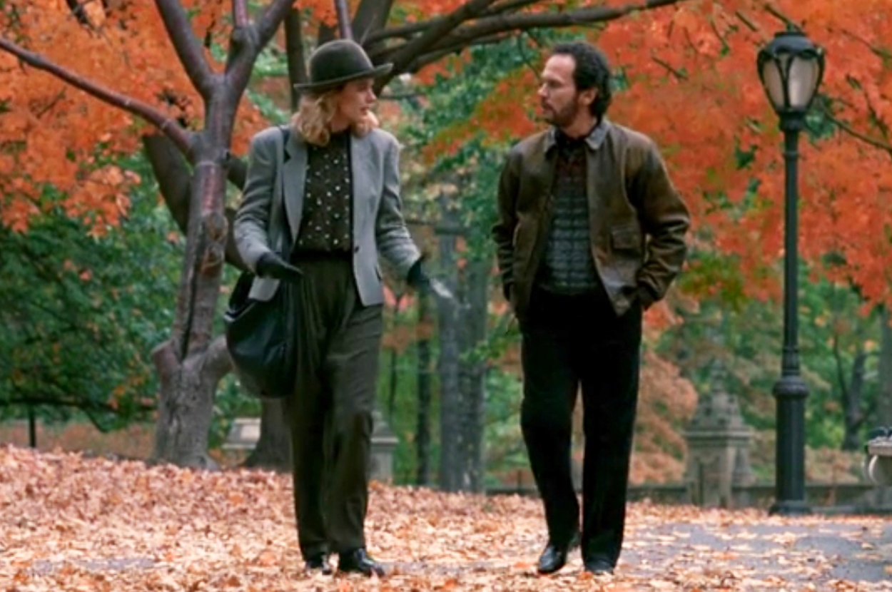 Sally and Harry from When Harry Mey Sally walk together through a park with autumn leaves on the ground and trees with colorful foliage