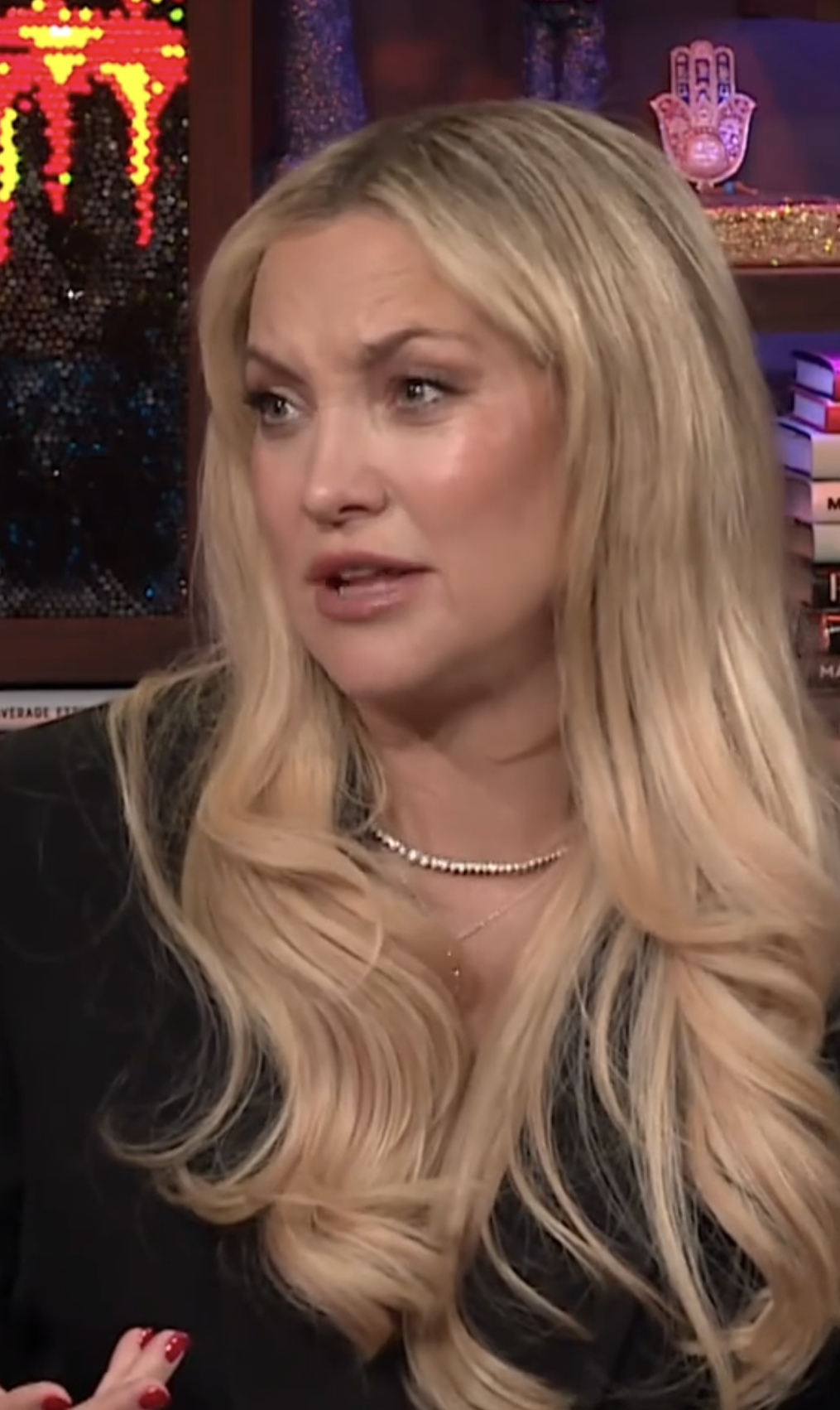 Kate Hudson is in a studio, wearing a black outfit and pearl necklace, speaking with an expressive gesture. Books and decorative items are in the background