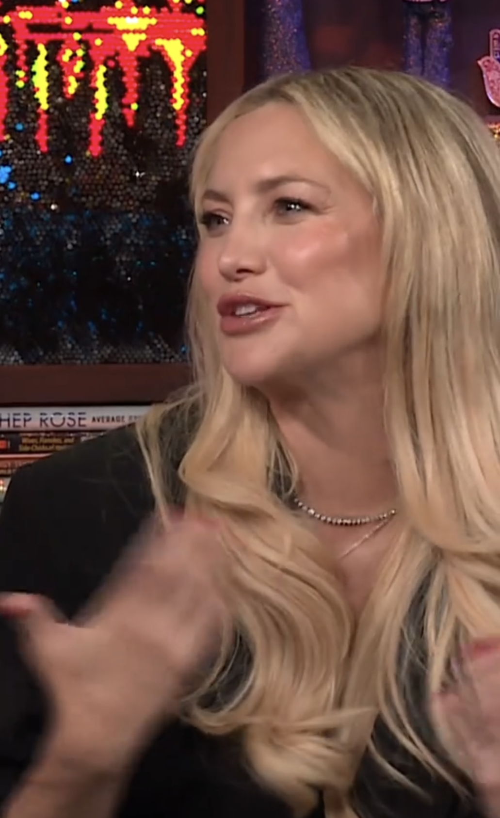Kate Hudson, dressed in elegant attire with long wavy blonde hair, gestures while speaking on a talk show set with colorful lights in the background