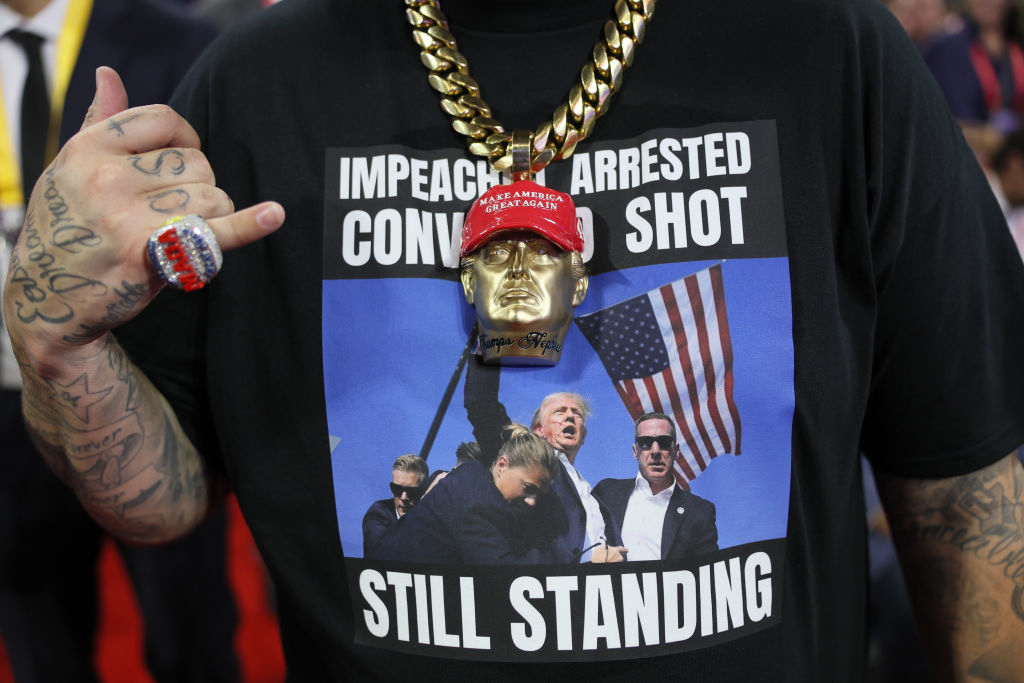Person wears a shirt reading &quot;Impeached, Arrested, Convicted, Shot, Still Standing&quot; with a picture of Donald Trump. They wear gold chains and rings