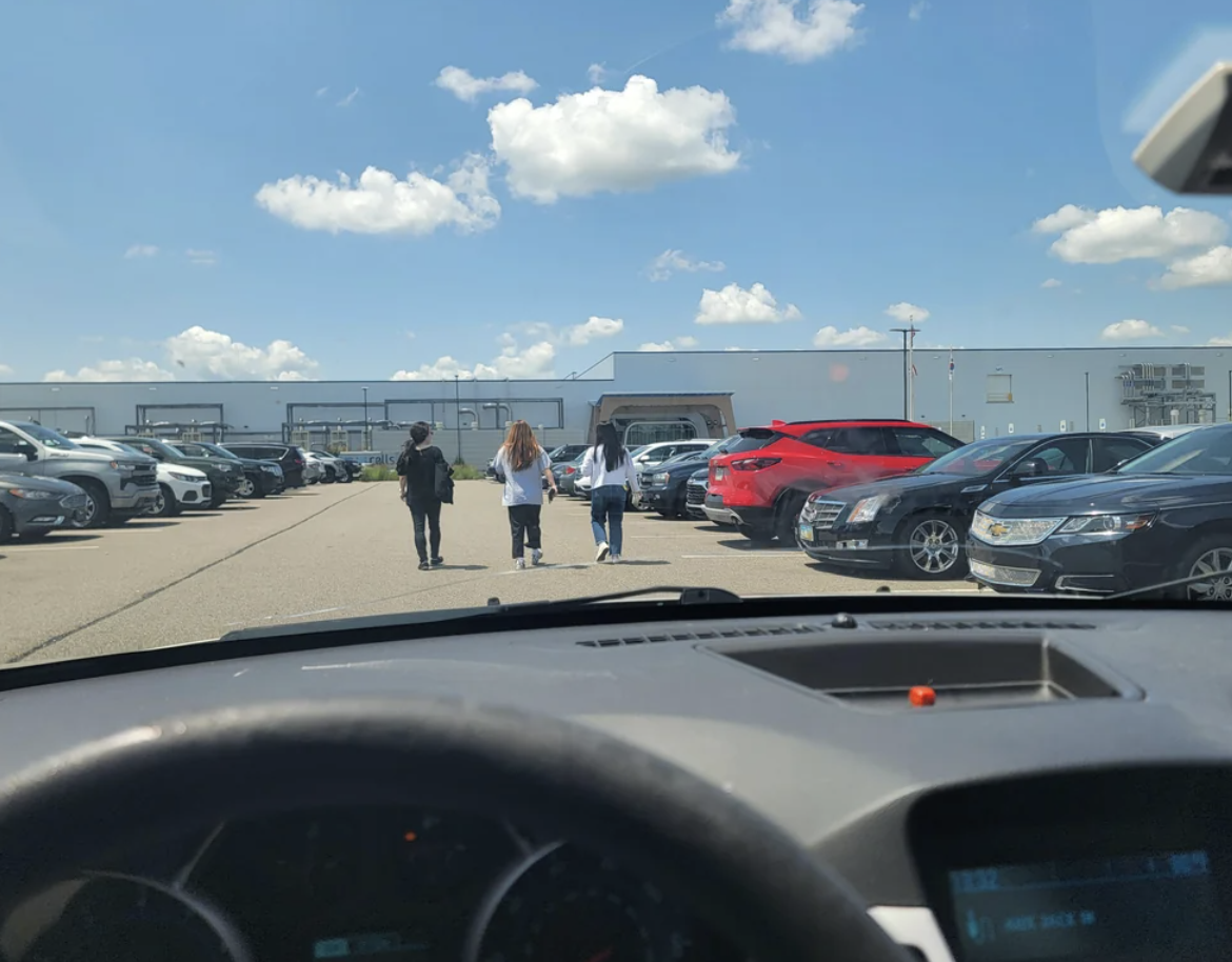 Three people walk towards a large industrial building through a parking lot filled with cars, viewed from inside a vehicle
