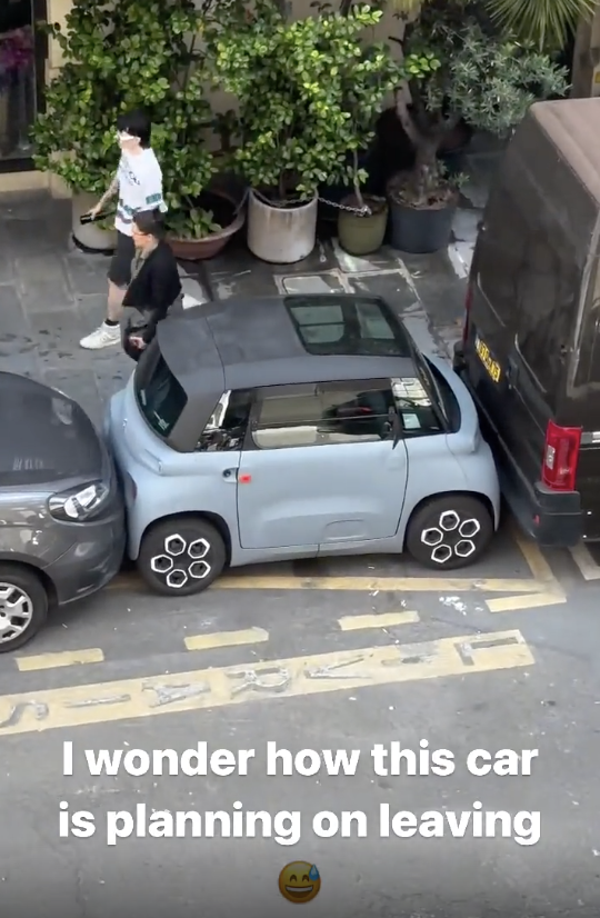 An extremely small car is tightly parallel parked between two larger vehicles on a street. Text overlay reads, "I wonder how this car is planning on leaving" with a smiley face emoji