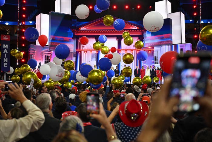 A crowd celebrates in front of a stage, surrounded by red, white, and gold balloons. The event has a festive atmosphere with many people taking photos