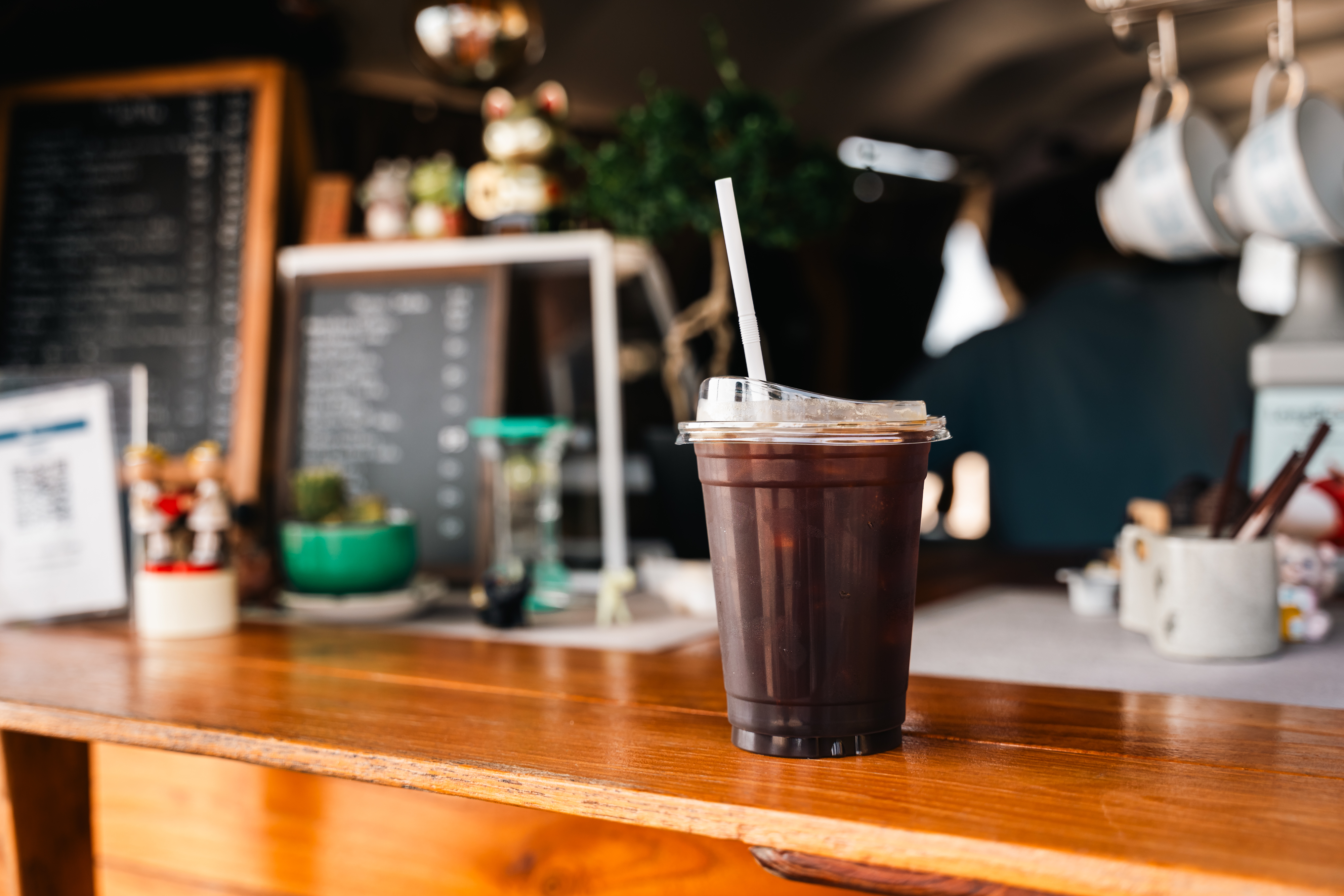 An iced coffee sits on a wooden counter in a cozy café, with a blurred menu and some decorations visible in the background