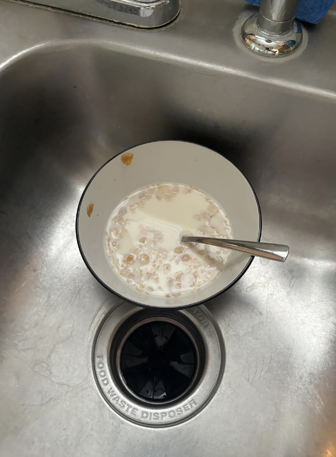 A bowl with partially eaten cereal and milk, placed in a stainless steel sink with a spoon in it. A food waste disposer is visible beneath the bowl