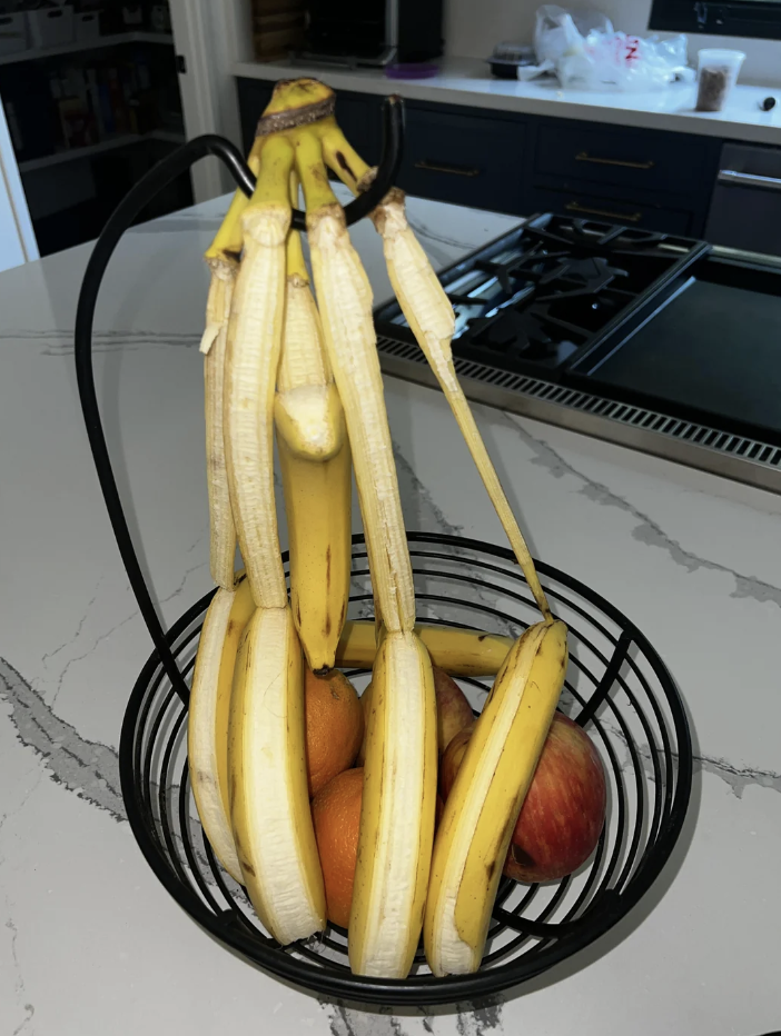 A banana holder with several peeled bananas, some oranges, and an apple inside a black wire basket on a kitchen counter