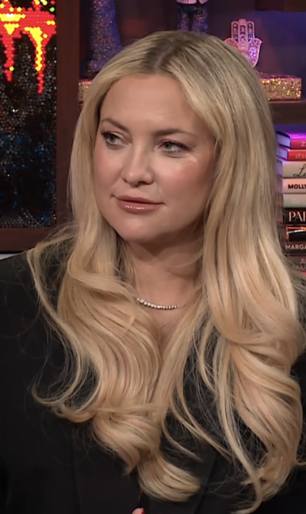 Kate Hudson with long flowing hair wearing a dark blazer and a pearl necklace in an indoor setting with books and decorative items in the background