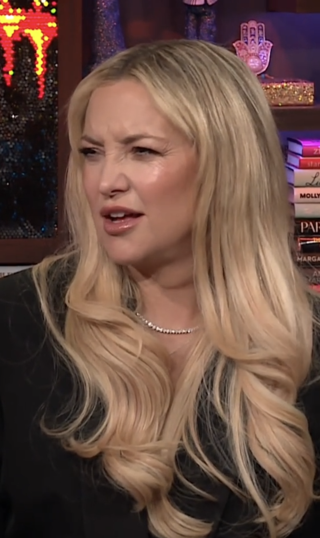 Kate Hudson appears confused during a talk show interview, wearing a black outfit and a pearl necklace. The background features books and decorative items