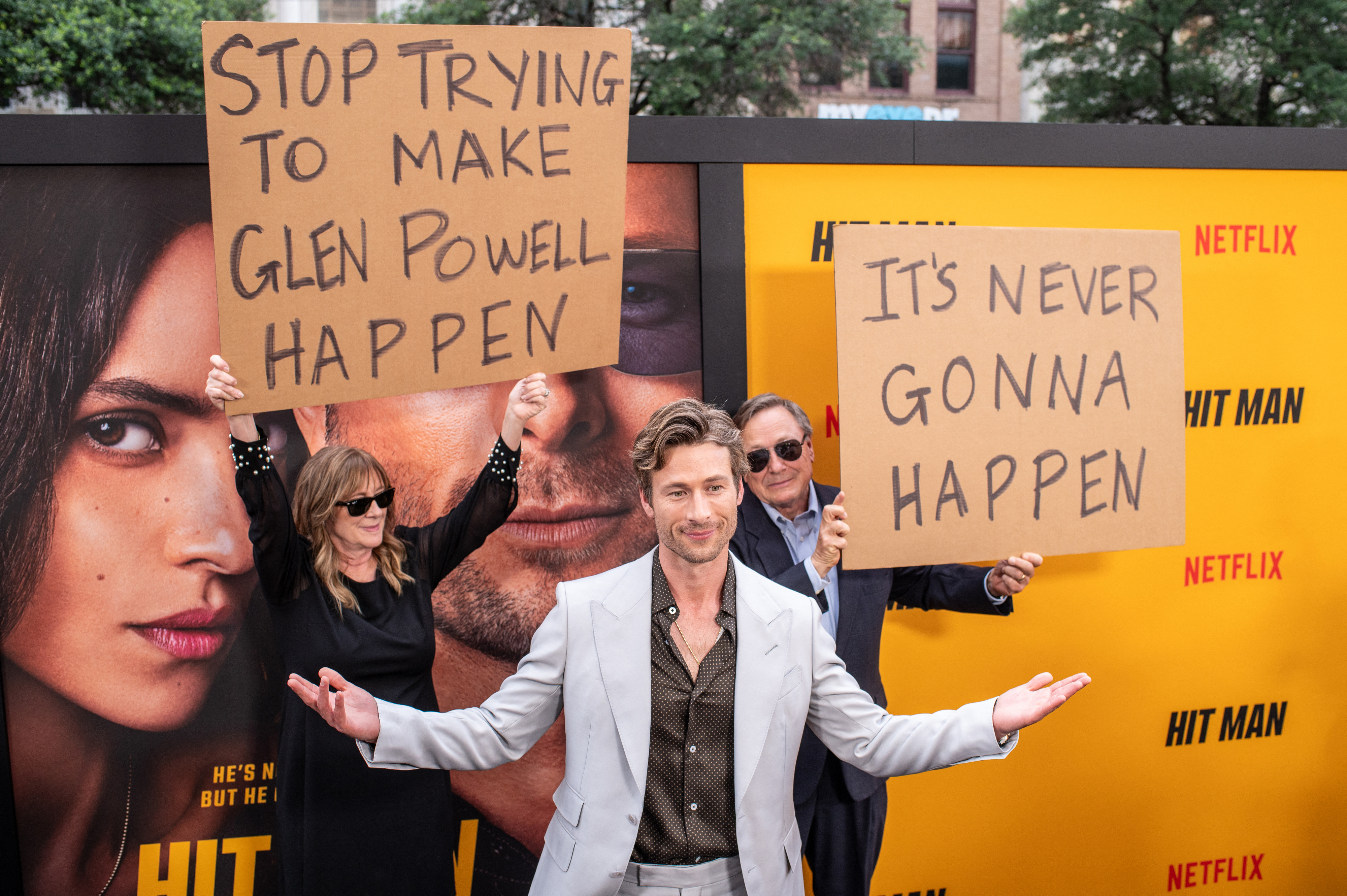 Glen Powell poses in a light suit with open arms. Behind him, his parents hold signs that say "Stop Trying To Make Glen Powell Happen" and "It's Never Gonna Happen"