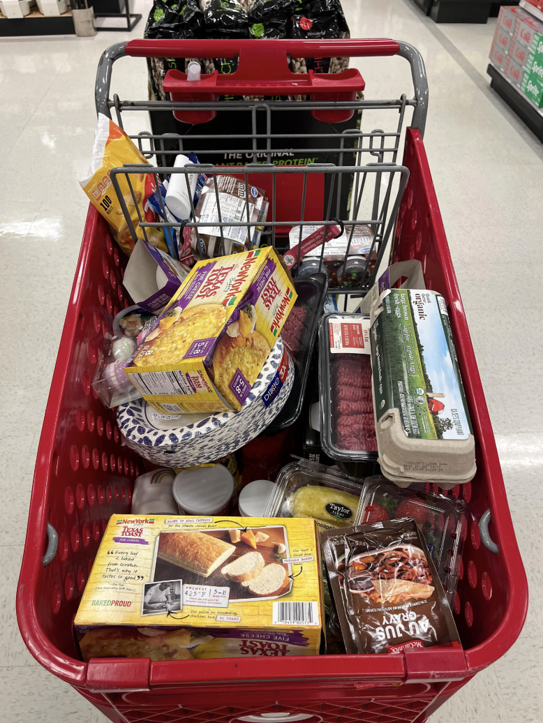 Shopping cart filled with various groceries, including frozen breakfast sandwiches, ground beef, eggs, bread, pineapples, and snacks, inside a store