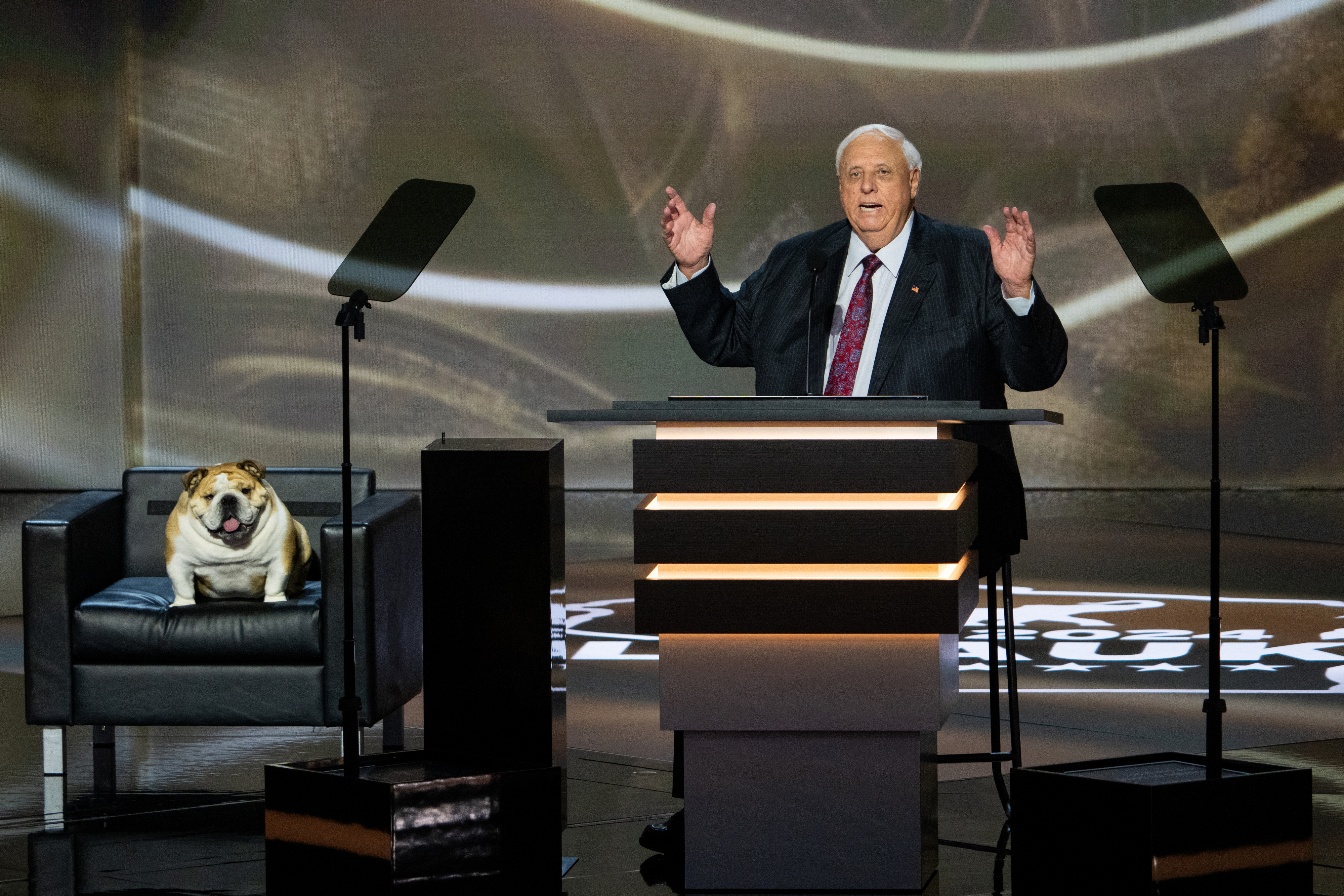 A man in a suit and tie speaks from a podium with teleprompters on either side. A bulldog sits on a chair to his left
