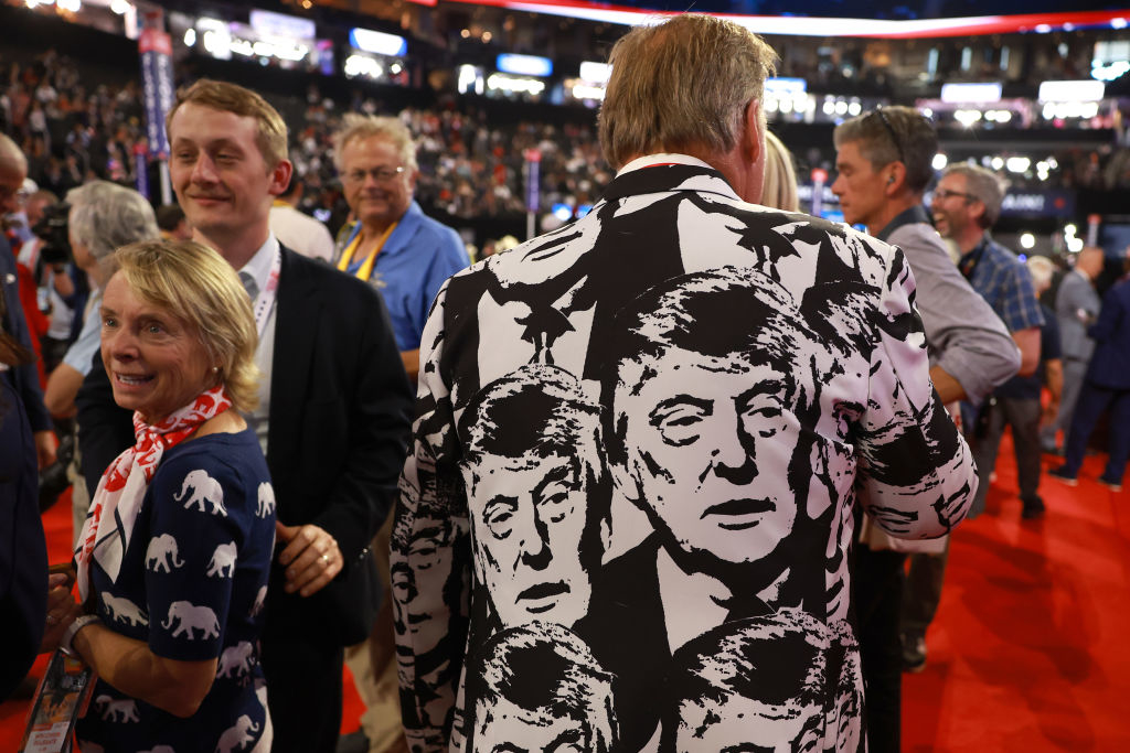 People at an event. A man is wearing a jacket with repeated prints of Donald Trump's face. Other people are engaged in conversation
