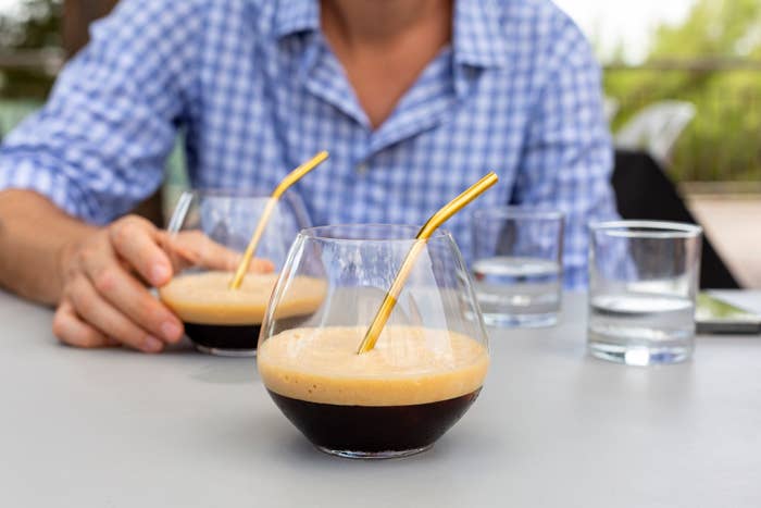 A person in a plaid shirt reaches for a glass of iced coffee with a gold straw. Another iced coffee glass and two water glasses are also on the table