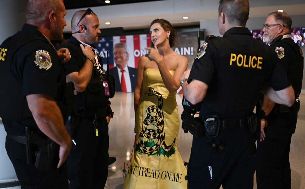 Joy Villa wears a &quot;Don't Tread on Me&quot; dress, speaking with five police officers inside a building with a crowd and a poster of Donald Trump in the background
