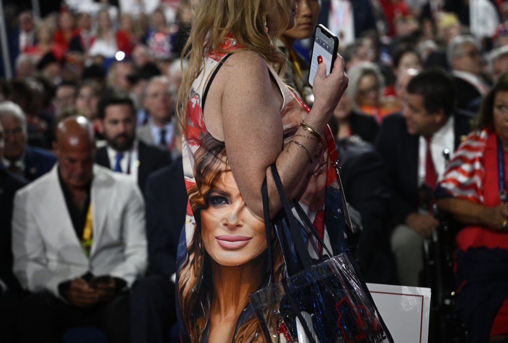A woman wears a sleeveless dress with an image of Melania Trump on it, holding her phone and shopping bag, among a crowd of seated people