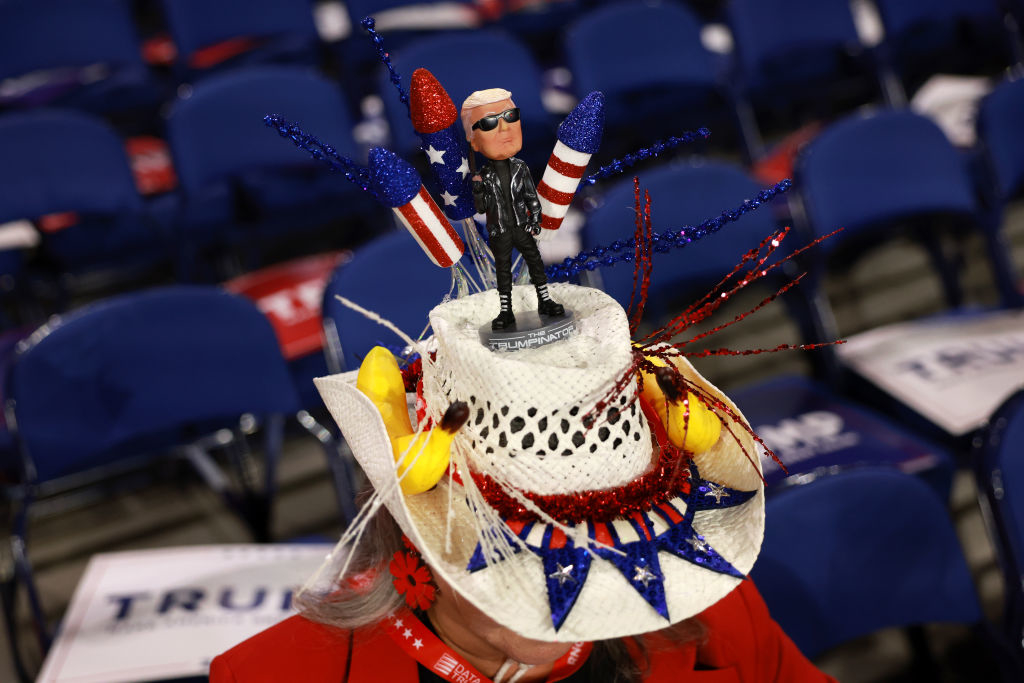 A person wears a festive hat featuring a bobblehead doll with American flag-themed accessories and decorations at a political event