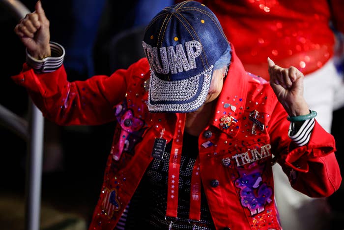 A person wearing a bedazzled &quot;TRUMP&quot; hat and a decorated red jacket celebrates with both fists in the air