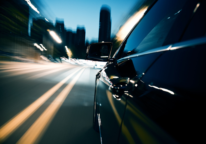 Close-up of a car driving fast on a city street at night, with blurred lights and buildings, emphasizing speed and motion. No people are visible