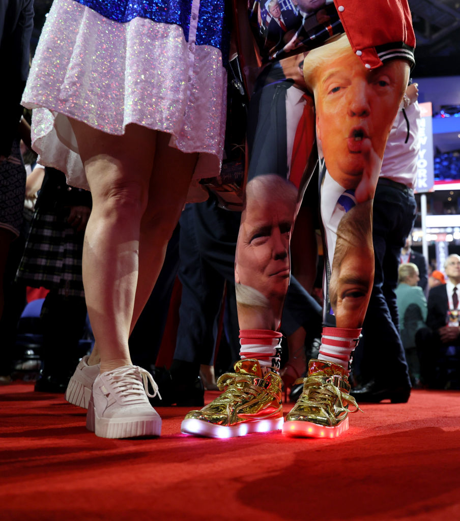 Two individuals stand on a red carpet. One wears white sneakers and a sparkly dress; the other wears gold light-up shoes and leggings printed with images of Donald Trump