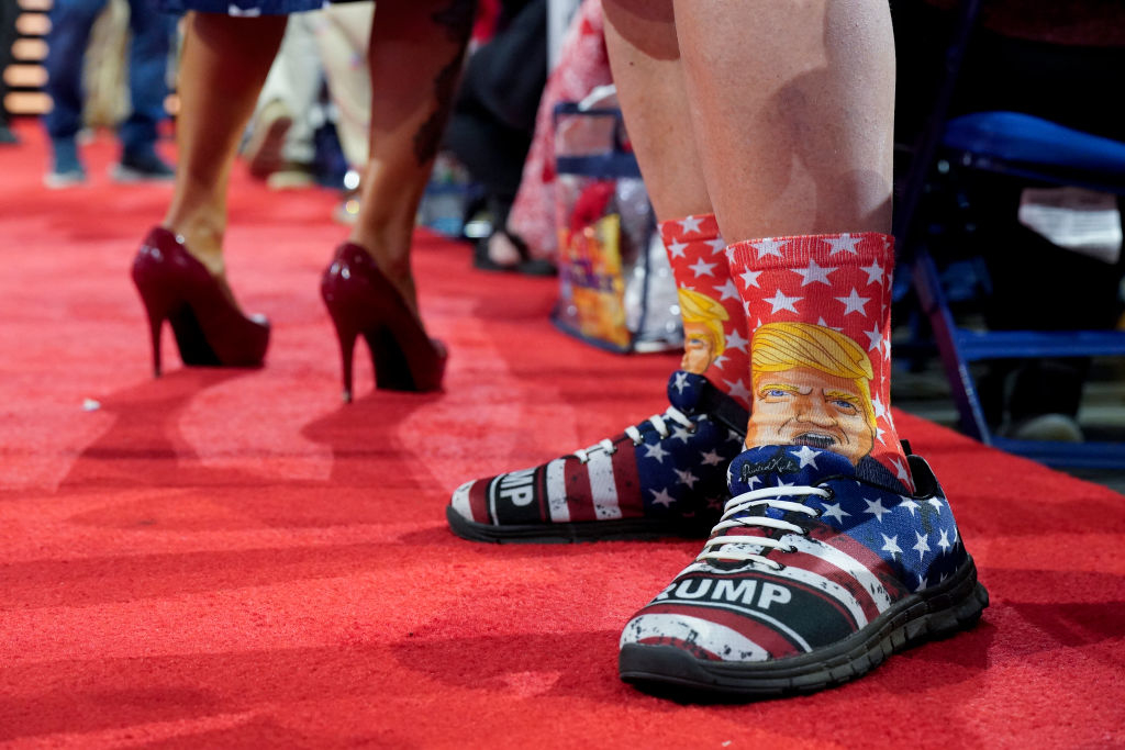Close-up of a person's feet wearing socks with Donald Trump's face and shoes with the word &quot;TRUMP&quot; on them. Another person in high heels is visible in the background