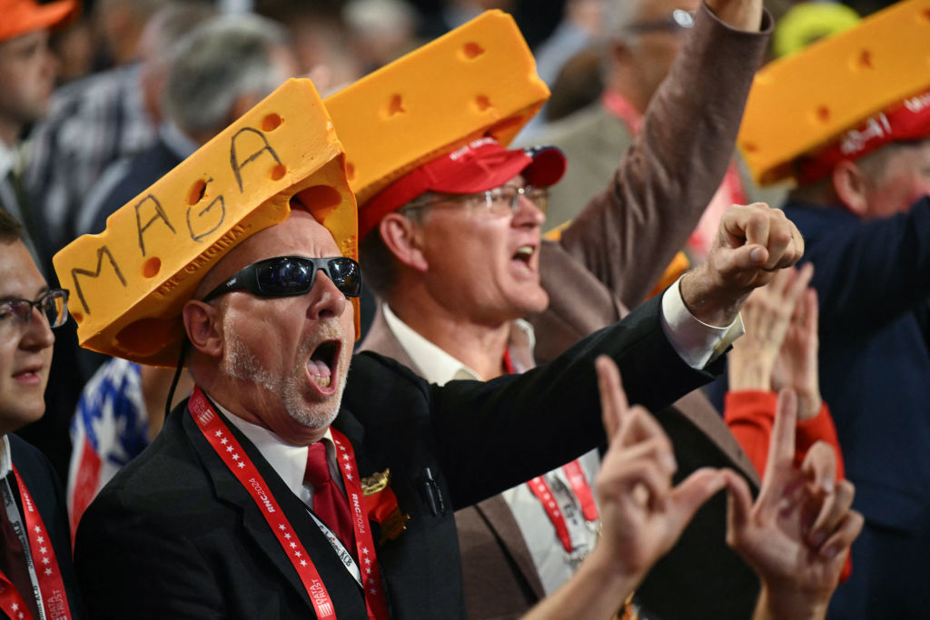 Supporters with cheese hats and &quot;MAGA&quot; text enthusiastically cheer at a rally, making hand gestures and wearing lanyards