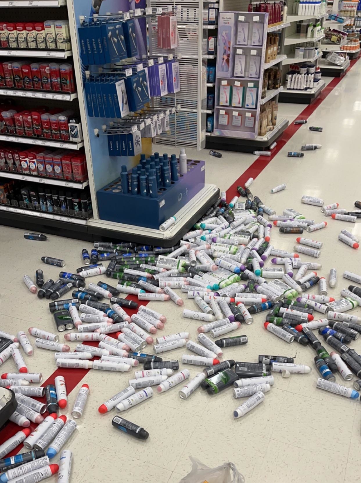 A store aisle with numerous scattered cans and bottles on the floor from a fallen shelf display. Various product types are visible on surrounding shelves