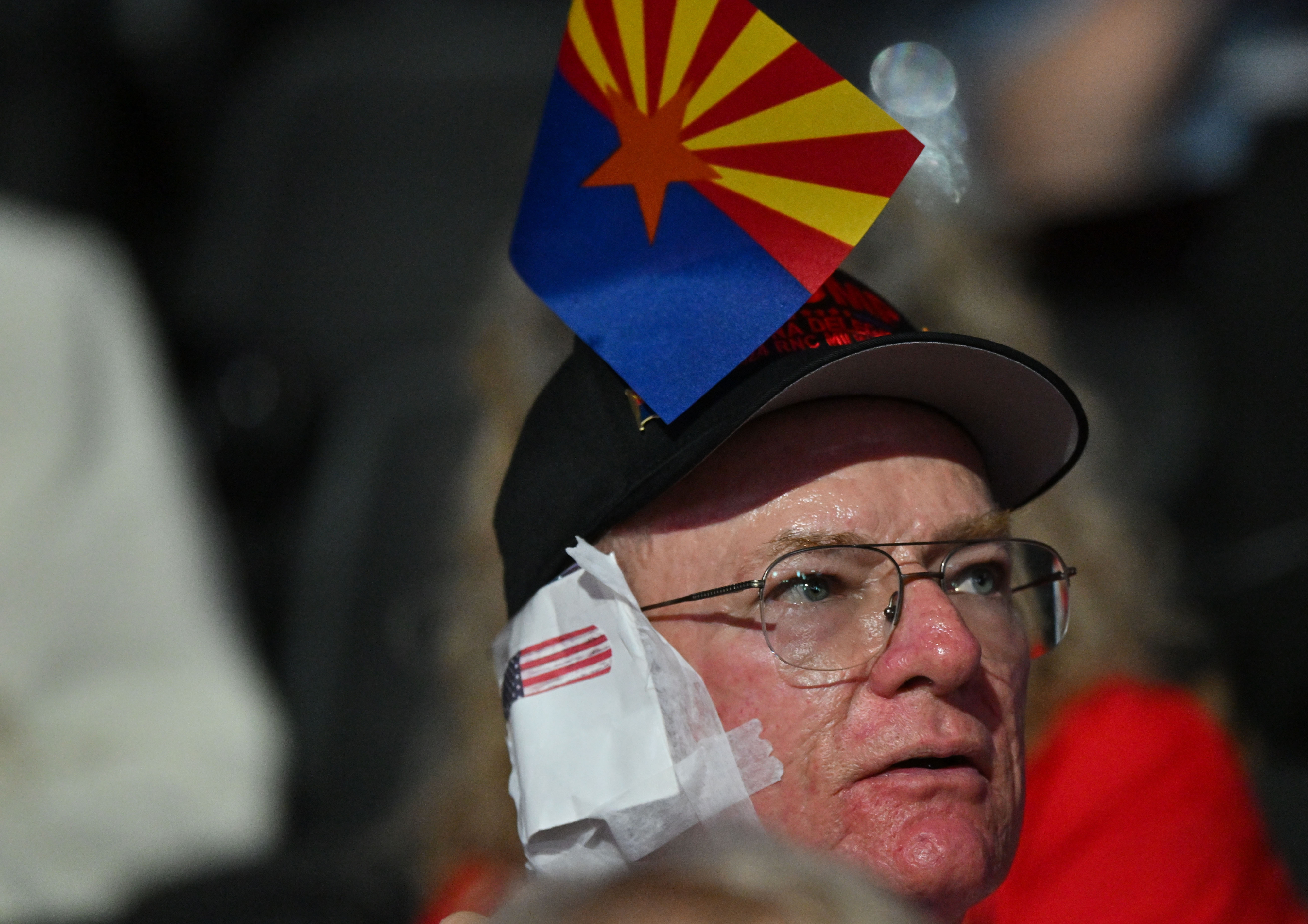 An older man is wearing a hat with the Arizona state flag and an American flag napkin under the brim. He appears to be at a public event