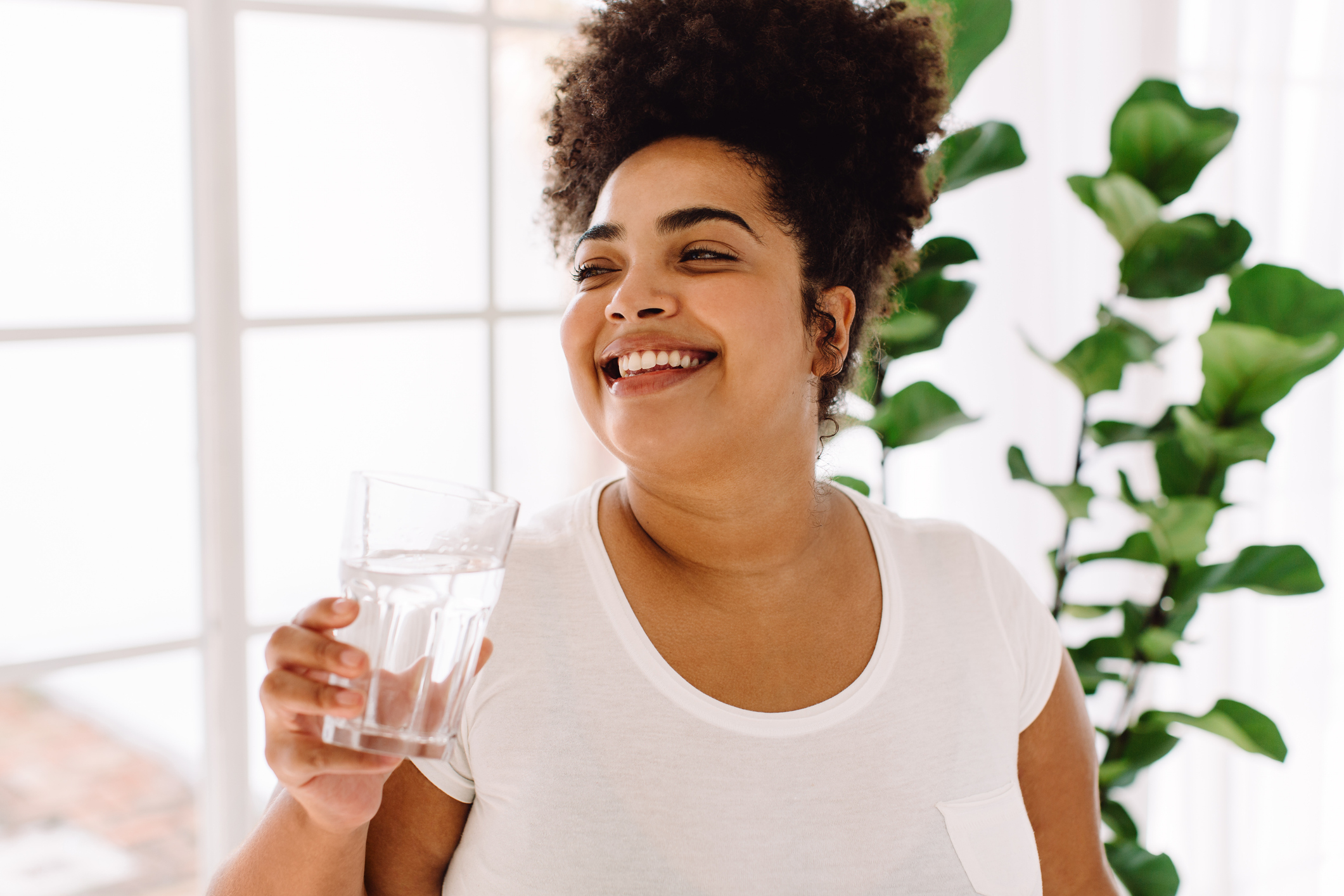 A woman with curly hair smiles while holding a glass of water, standing in a bright room with plants in the background
