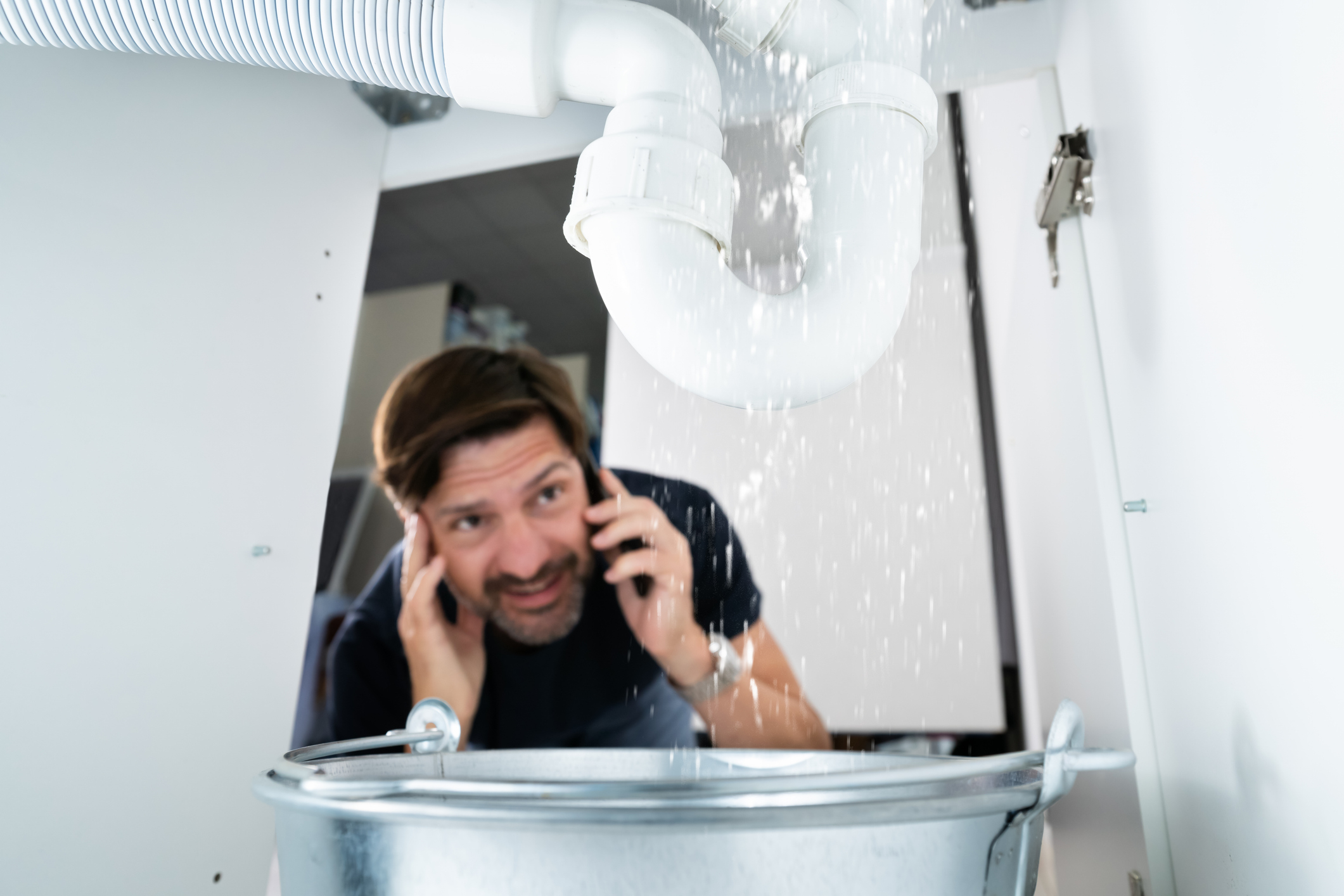 A man talking on the phone is looking at a leaking pipe under a sink, with water spilling into a metal bucket