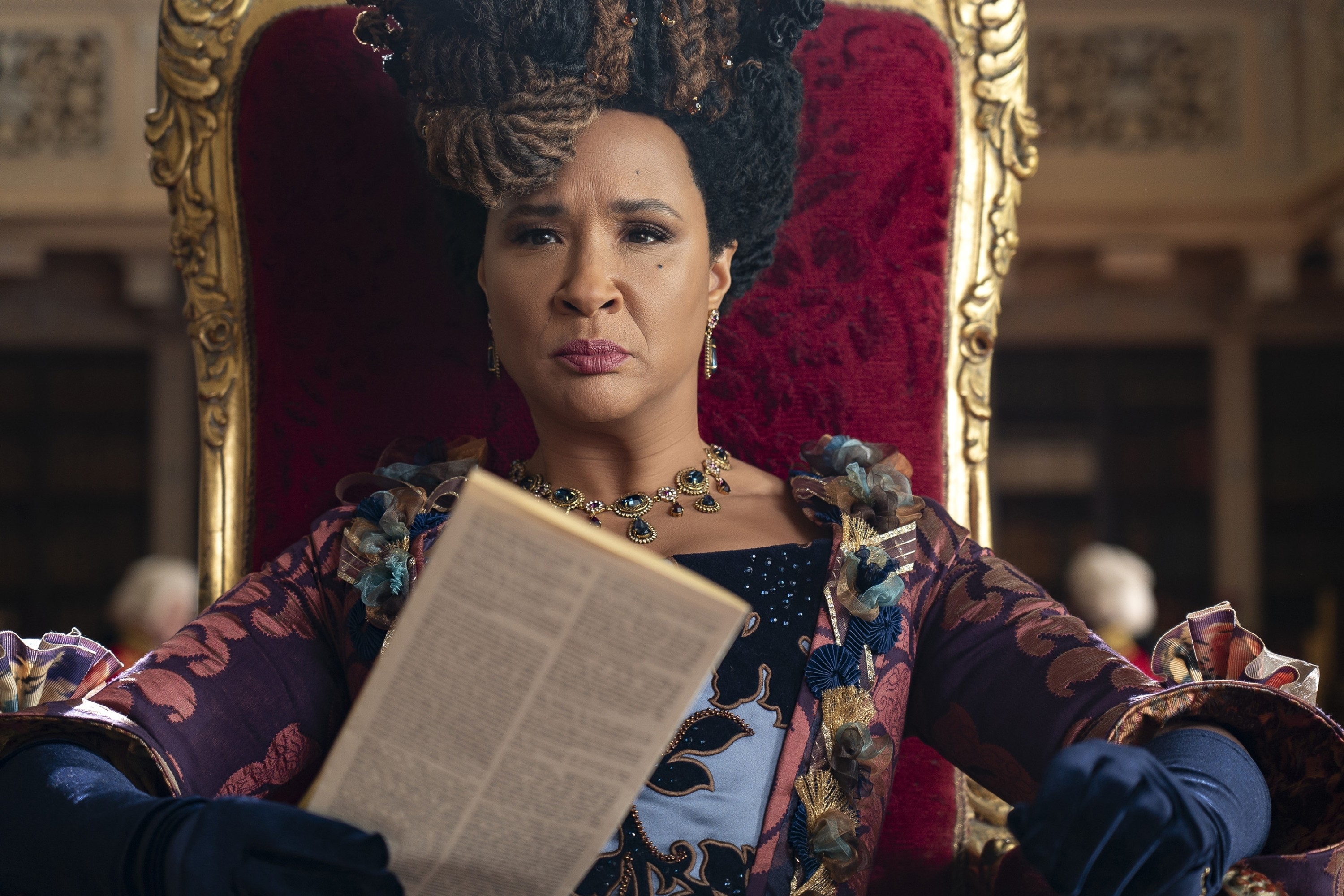 Golda Rosheuvel dressed in regal, period attire with elaborate hair, sits on a throne reading a document in a scene from TV show &quot;Bridgerton.&quot;