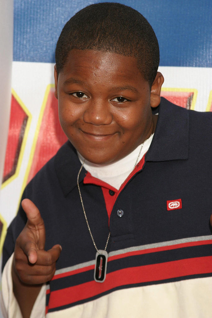 A person wearing a polo shirt with red, white, and black stripes, and a necklace with a rectangular pendant, pointing towards the camera with both hands